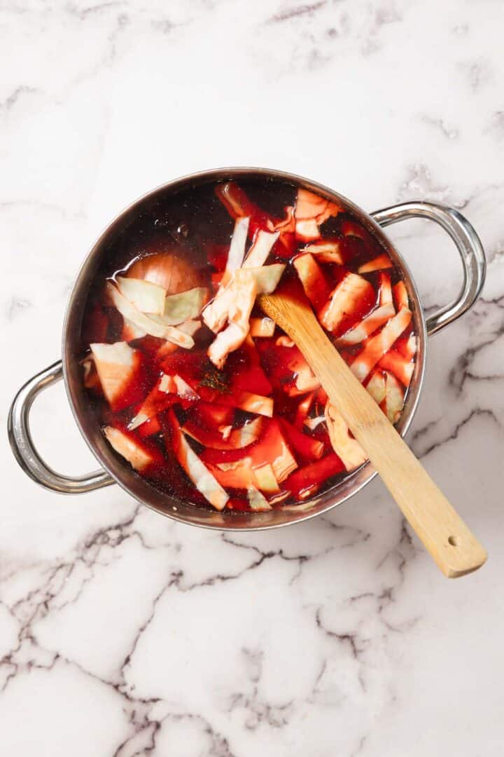 A pot of Chicken Borscht soup with cabbage and beets, stirred with a wooden spoon on a marble countertop.