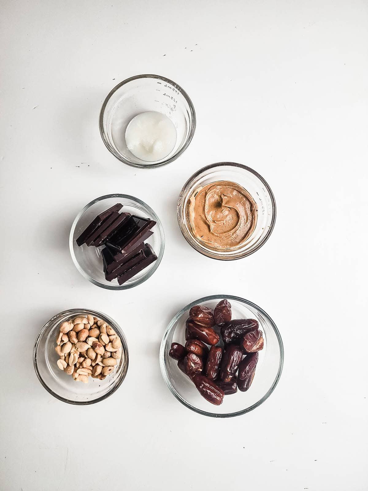 Five glass bowls with coconut oil, peanut butter, dark chocolate, peanuts, and dates on a white background.