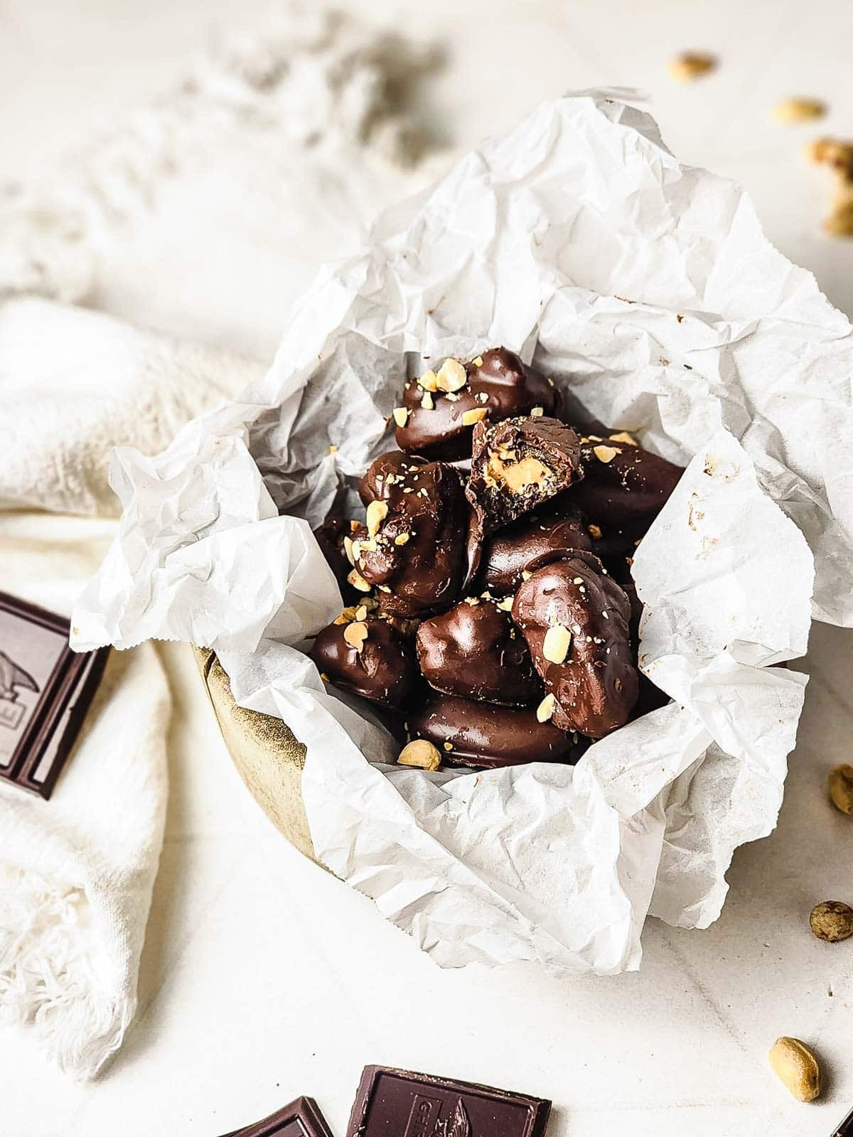 A bowl lined with parchment paper filled with chocolate-covered snacks, topped with chopped nuts.