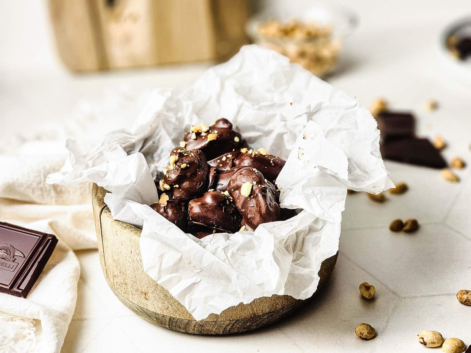 A wooden bowl lined with parchment paper holds chocolate-covered treats topped with chopped nuts.
