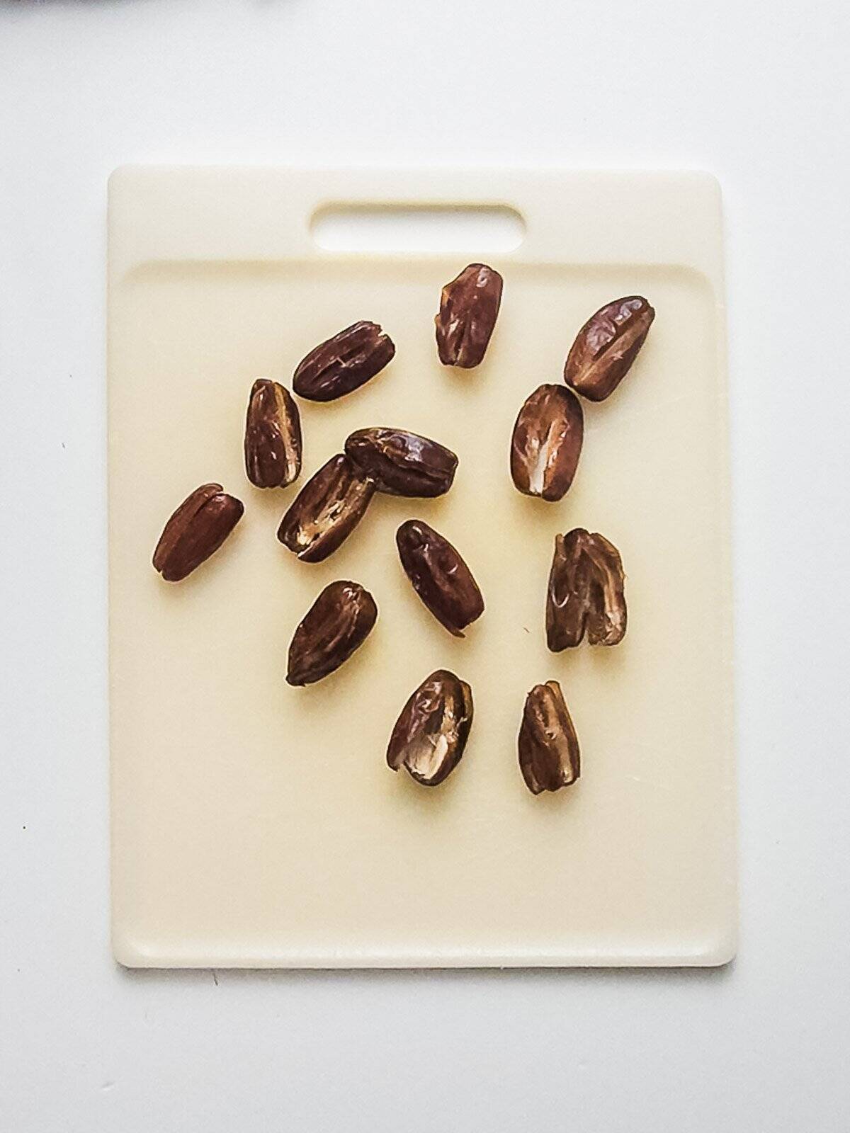 Twelve pitted dates are scattered on a white cutting board with a handle, on a white surface.