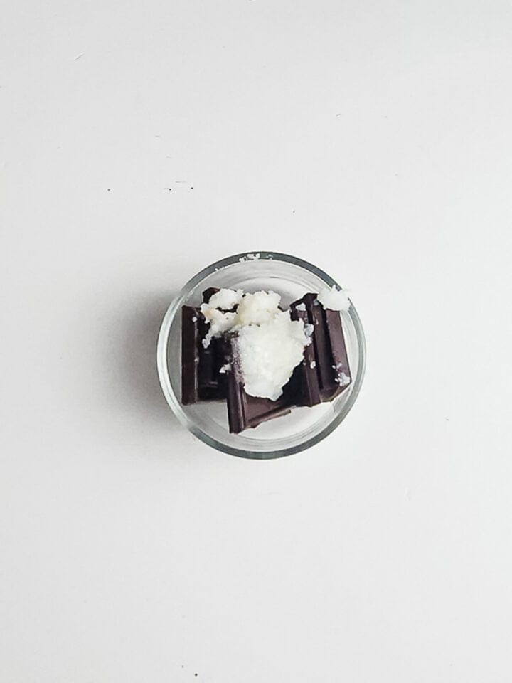 A glass bowl with dark chocolate pieces and a dollop of white coconut oil on a white background.