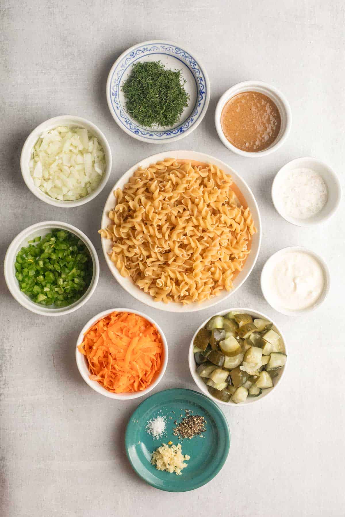 Nine bowls with ingredients for Pickle Pasta Salad, including pasta, chopped veggies, tangy pickles, seasonings, and dressing.