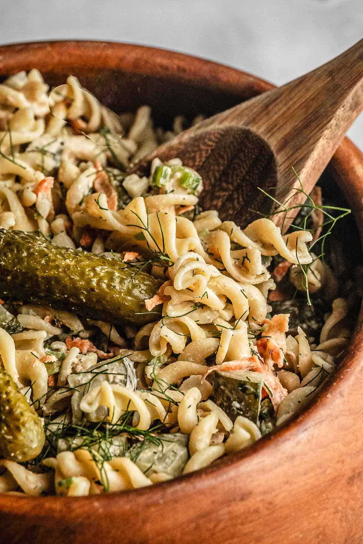 Rotini pasta salad with dill, pickles, and herbs in a wooden bowl with a wooden spoon.