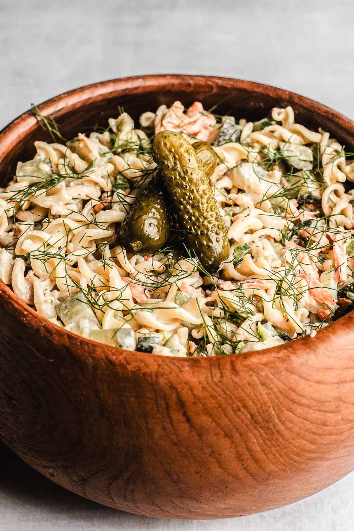 Wooden bowl filled with creamy Pickle Pasta Salad, garnished with fresh dill and two pickles on top.