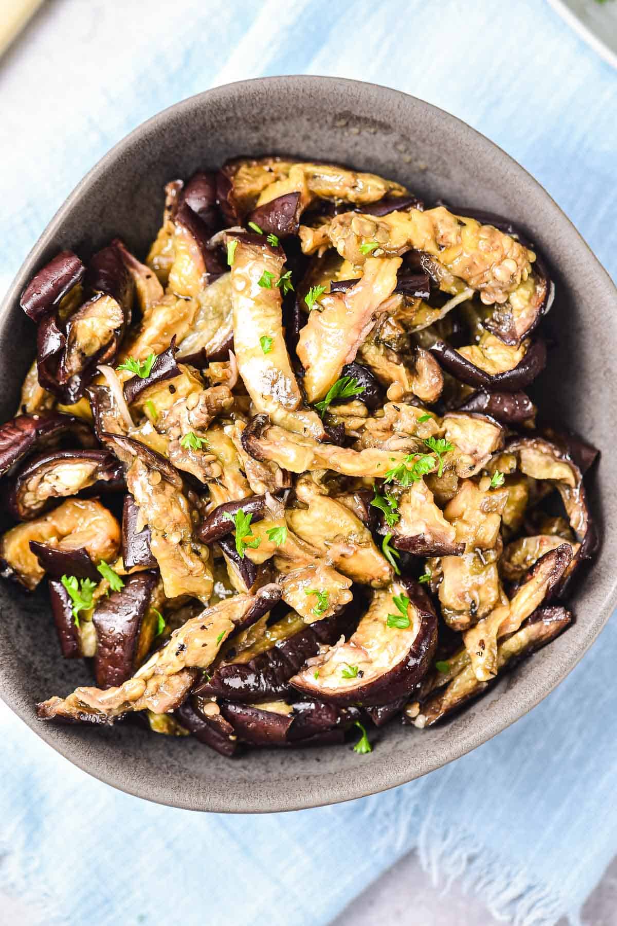 Sliced roasted eggplant garnished with chopped parsley in a dark bowl on a light blue cloth.