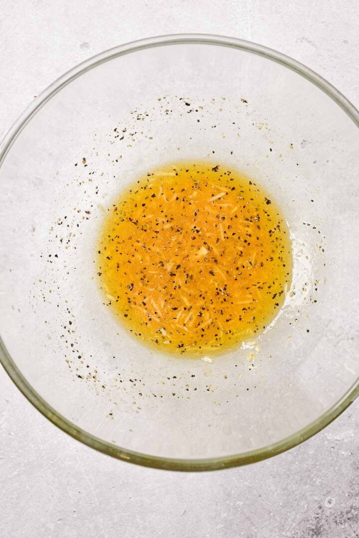 A clear glass bowl with a yellow vinaigrette dressing speckled with black pepper on a light surface.