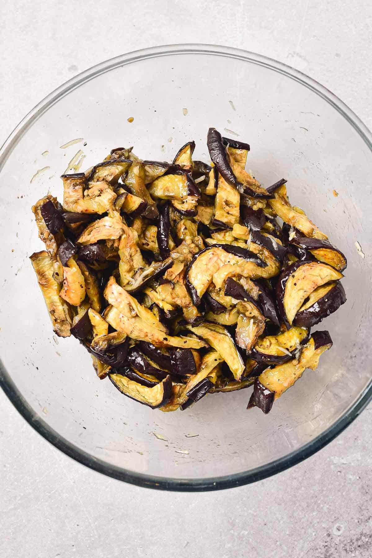 Sliced, roasted eggplant pieces in a clear glass bowl on a light-colored surface.