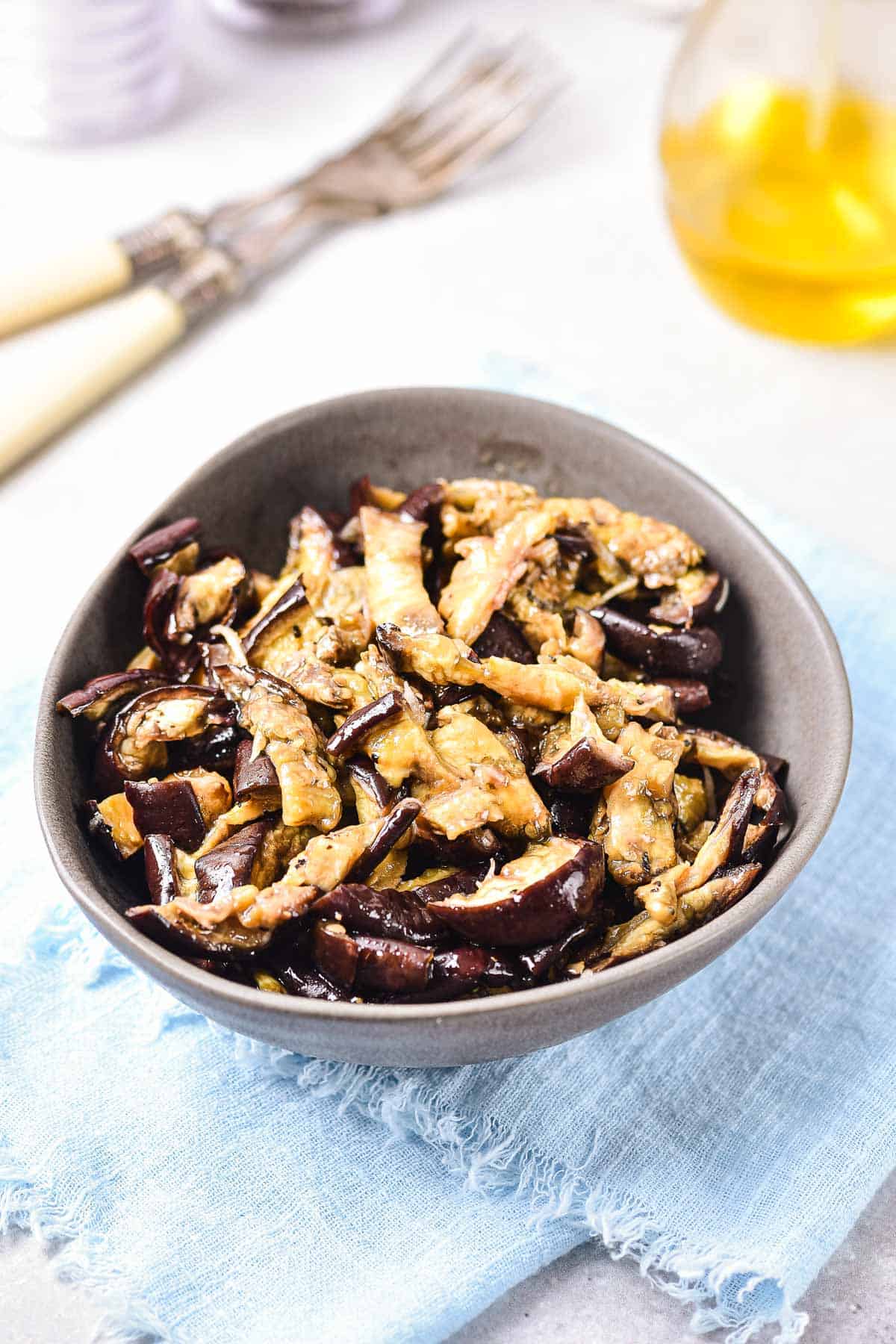 A bowl of cooked, sliced eggplant on a blue cloth with forks and a bottle of oil in the background.