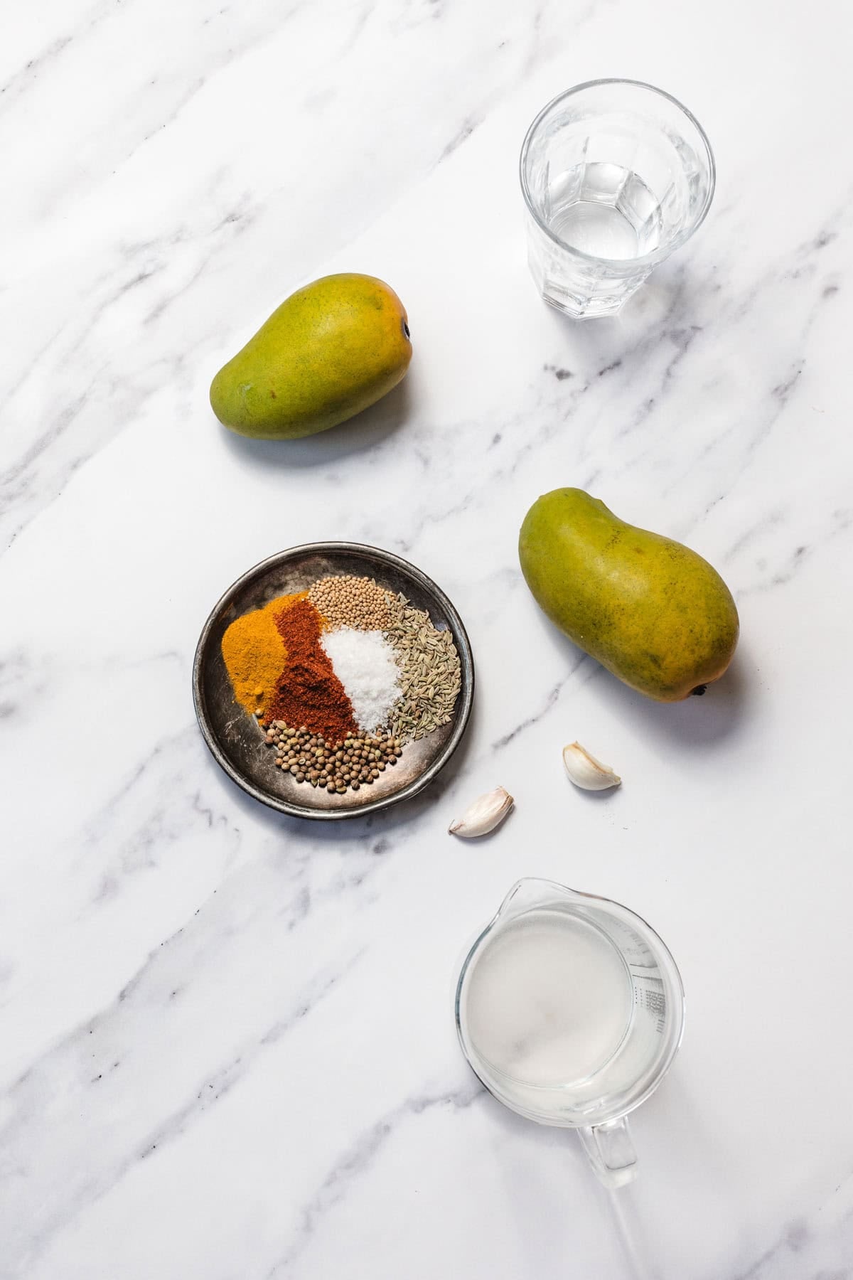Two mangoes, a plate of spices, two garlic cloves, a glass of water, and a jug of milk on a marble surface.