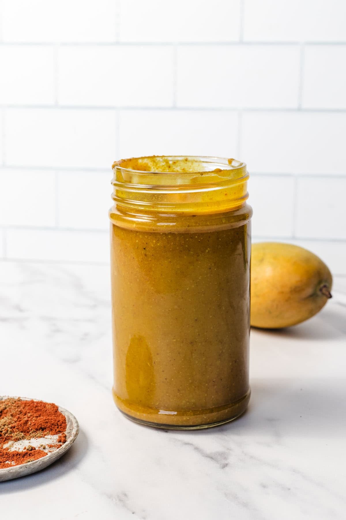 A jar of golden mango salad dressing on a marble counter, with a mango and spices nearby.