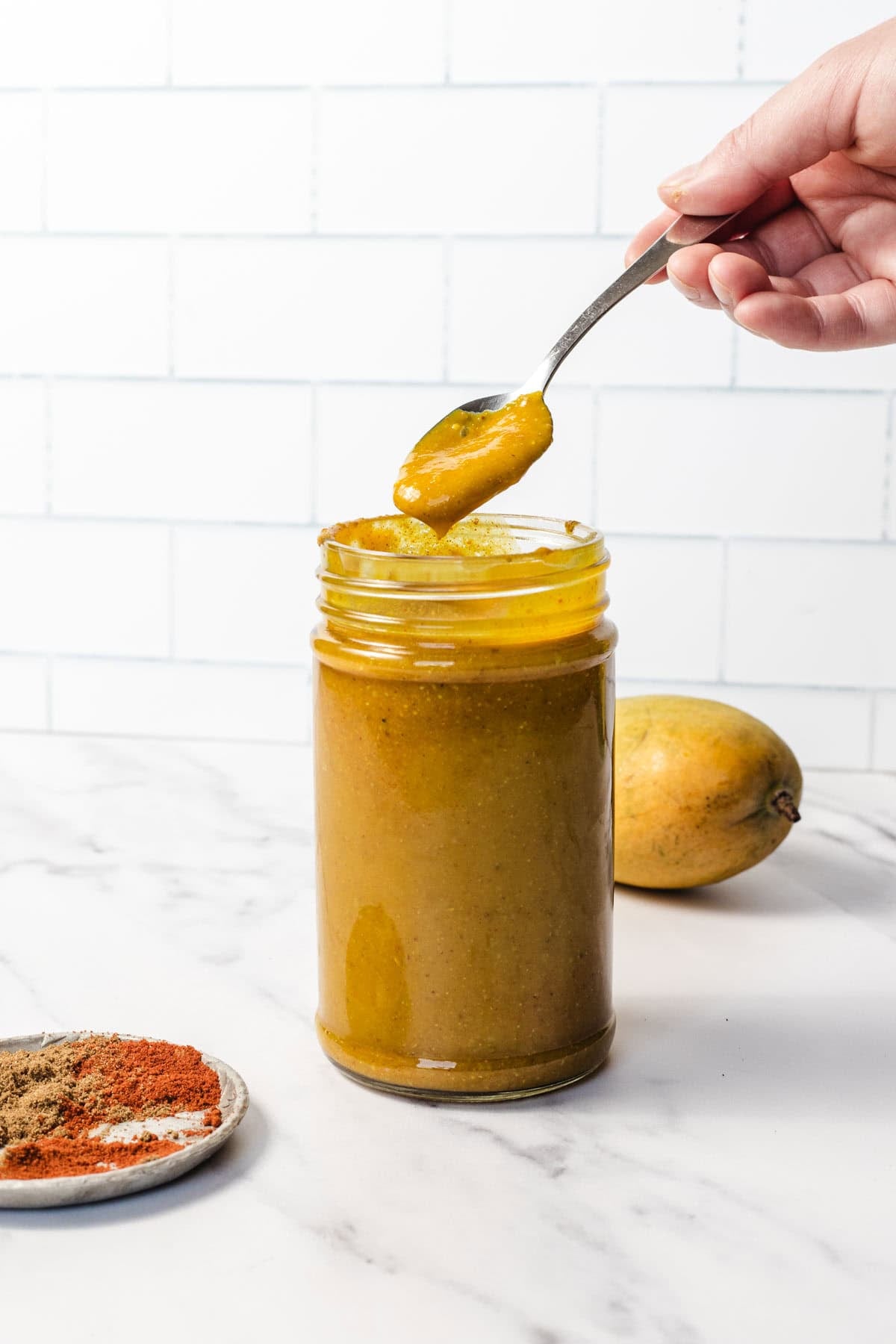 A hand holds a spoonful of yellow mango chutney above a jar; spices and a mango are nearby.