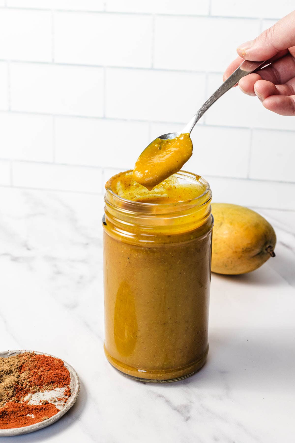 A hand holds a spoonful of yellow sauce above a jar, with spices and a mango on a white surface nearby.
