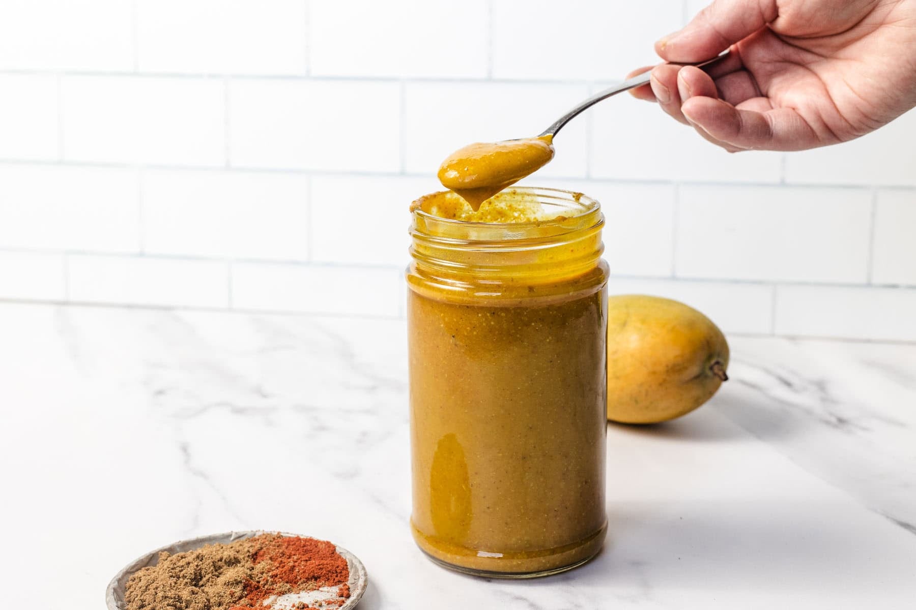 A hand holds a spoon of yellow sauce above a jar, with spices and a mango nearby on a white countertop.