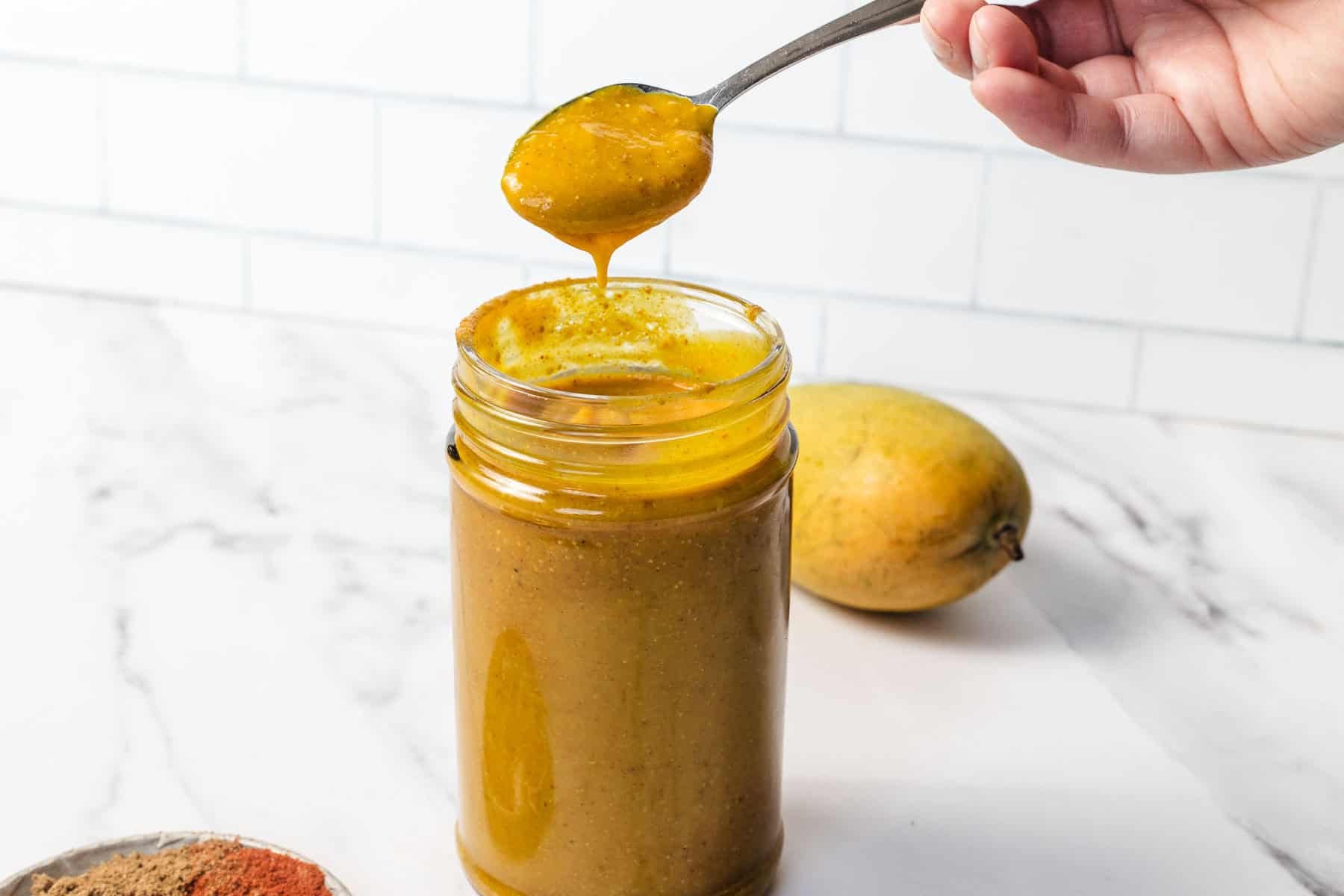 A jar of mango chutney with a spoon lifting some out; a fresh mango sits in the background.
