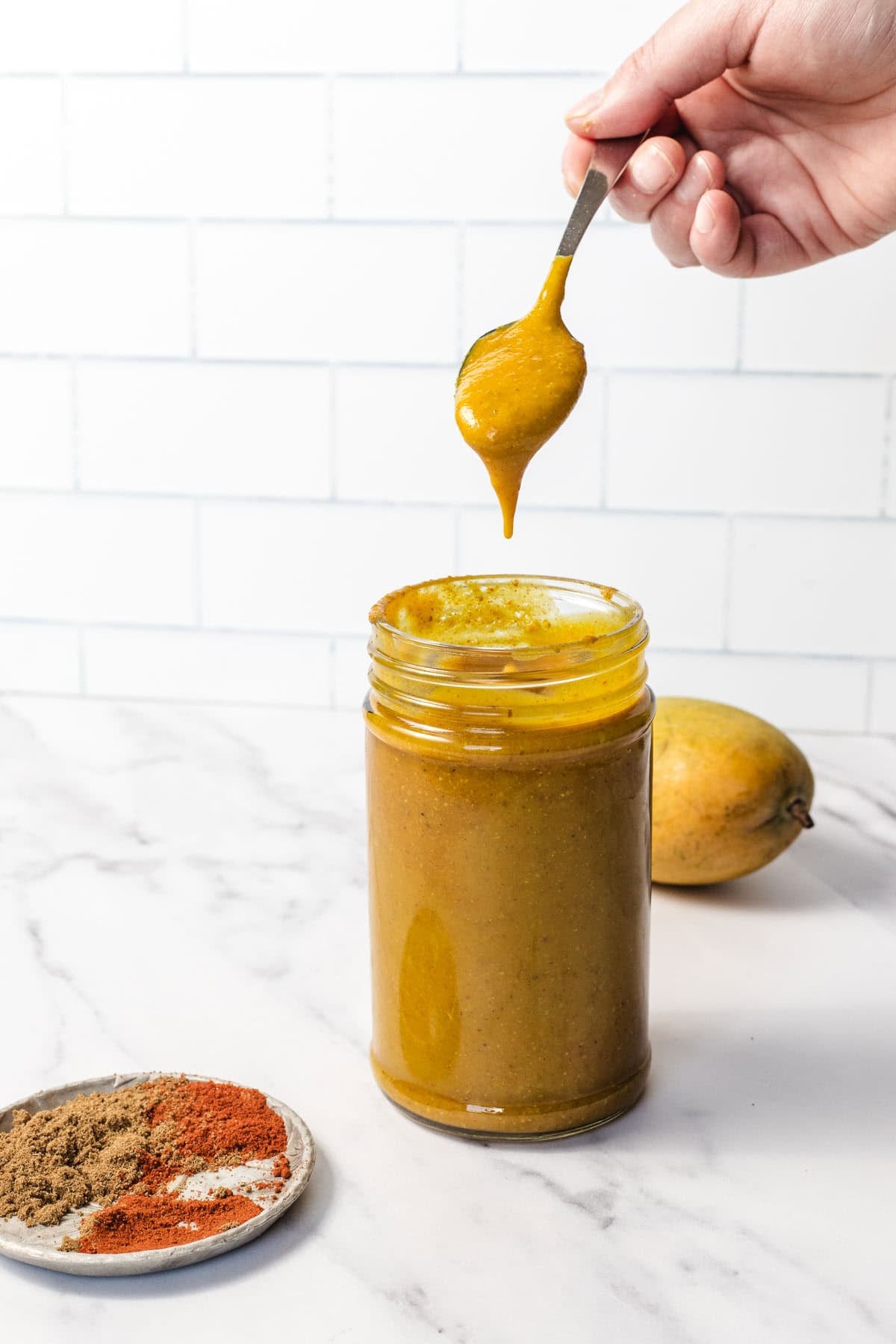 A hand drizzles yellow sauce from a spoon into a jar, with spices and a mango nearby on a marble counter.