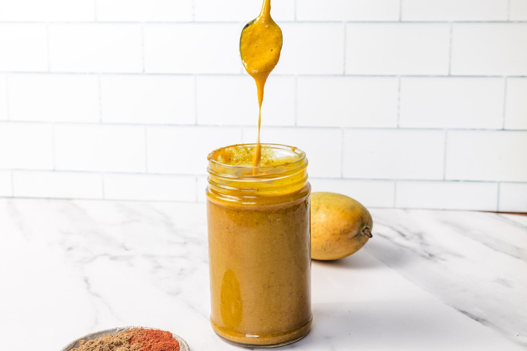 A spoon drizzles golden turmeric paste into a glass jar on a marble counter, with mango and spices nearby.