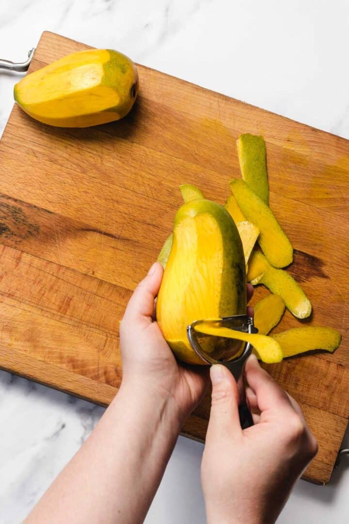 Hands peeling a yellow mango with a peeler on a wooden cutting board, with another mango nearby.