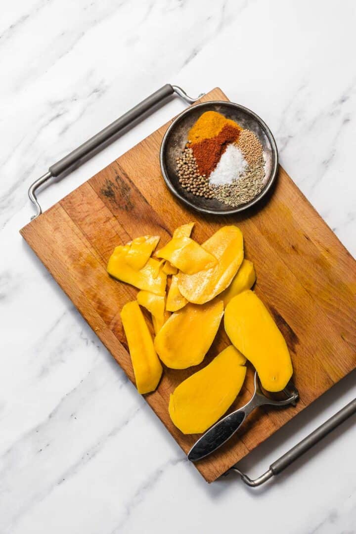 Sliced mango on a wooden cutting board with a bowl of assorted spices on a marble surface.