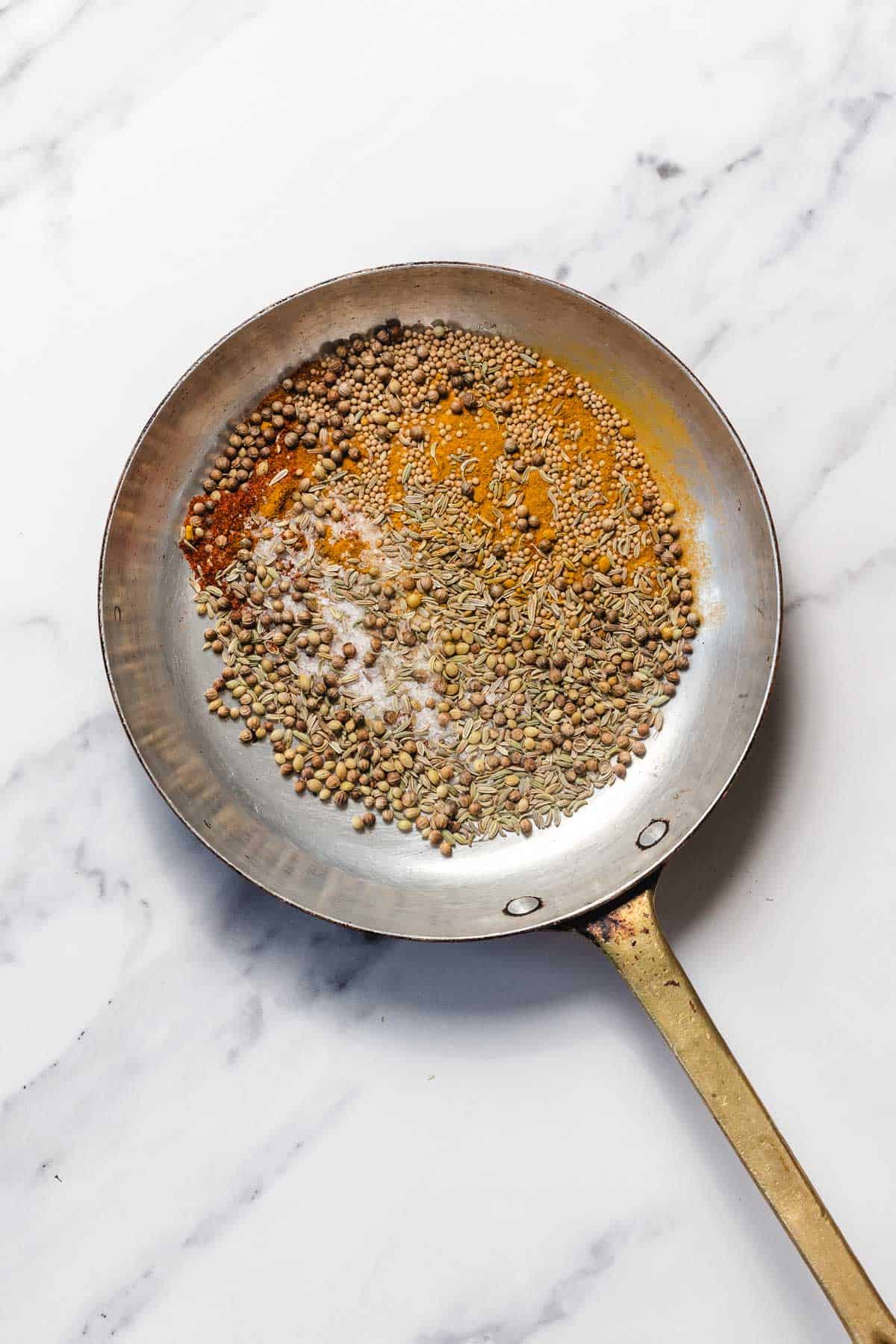 A metal pan with assorted whole spices and turmeric powder on a white marble surface.