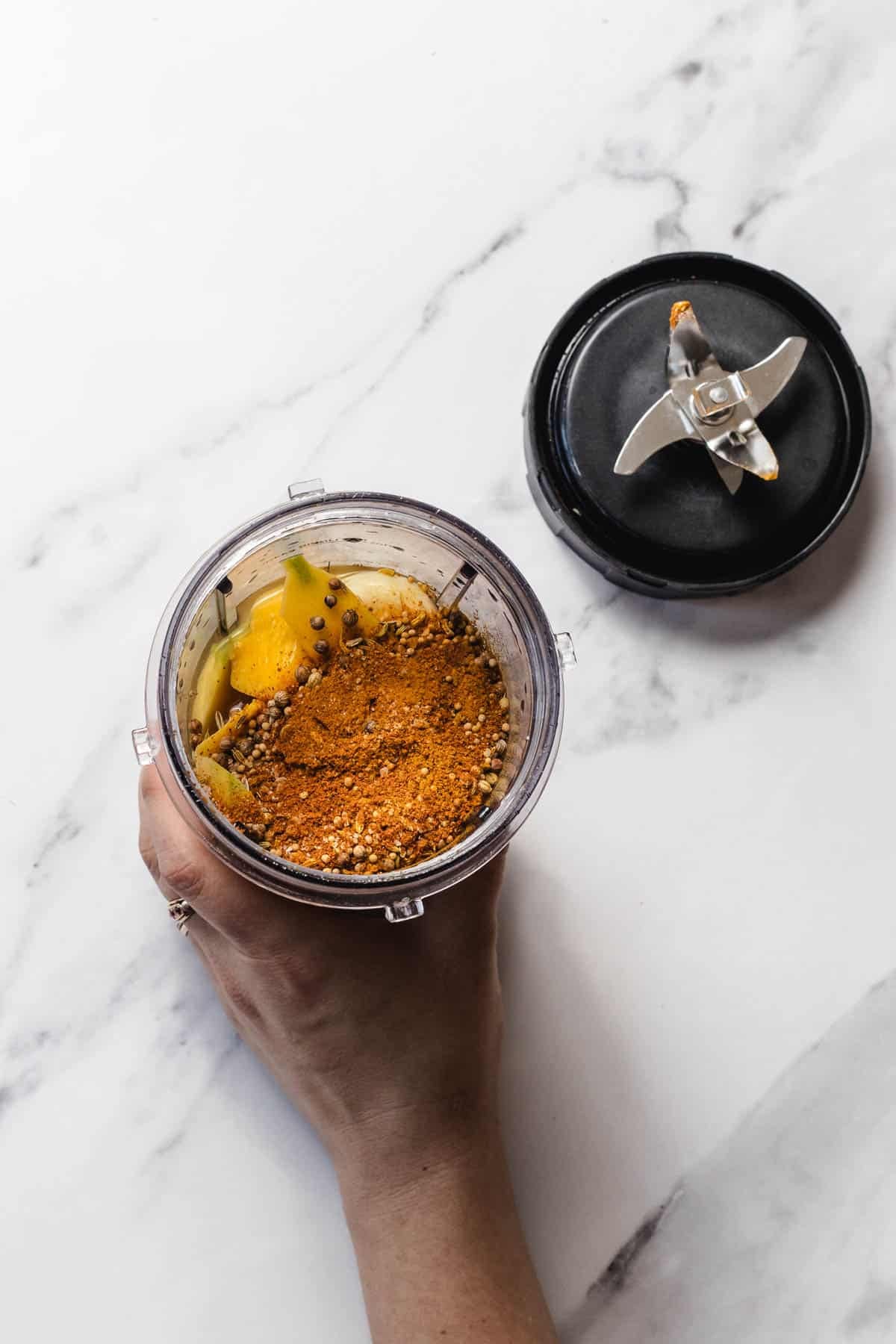 A hand holds a blender cup with spices and vegetables inside, next to a removed blade lid on a white surface.