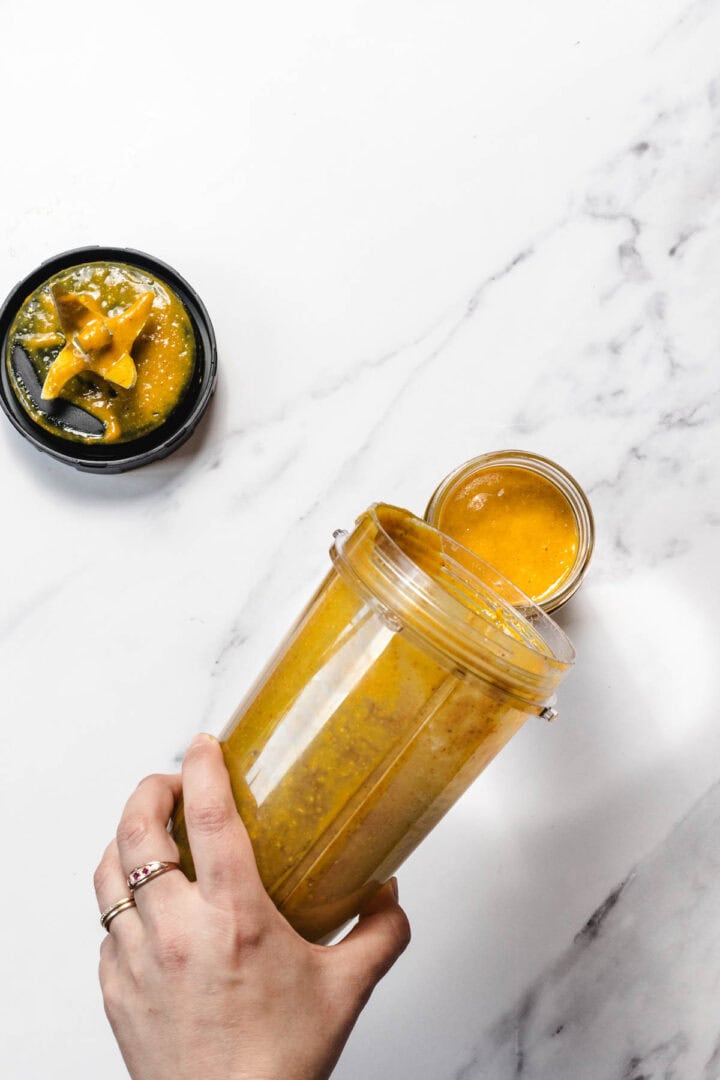 A hand holds a blender cup pouring yellow sauce into a small glass jar on a marble surface.