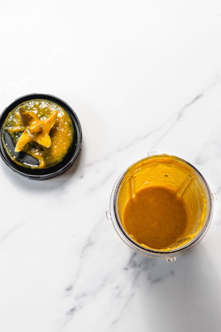A blender cup with orange sauce inside and a messy blender lid on a white marble surface.