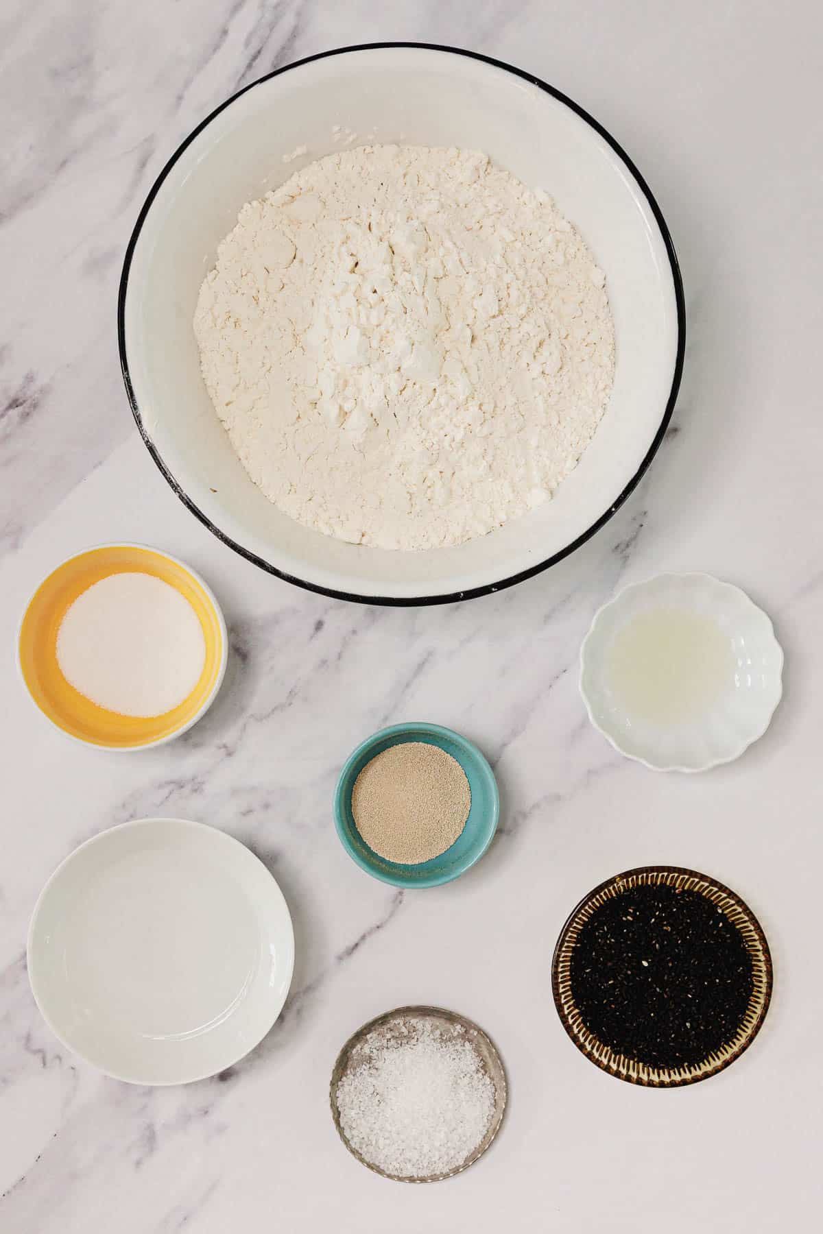 A bowl of flour with small dishes of yeast, salt, oil, sugar, water, and black sesame seeds on a marble surface—everything you need for an Easy Challah recipe.
