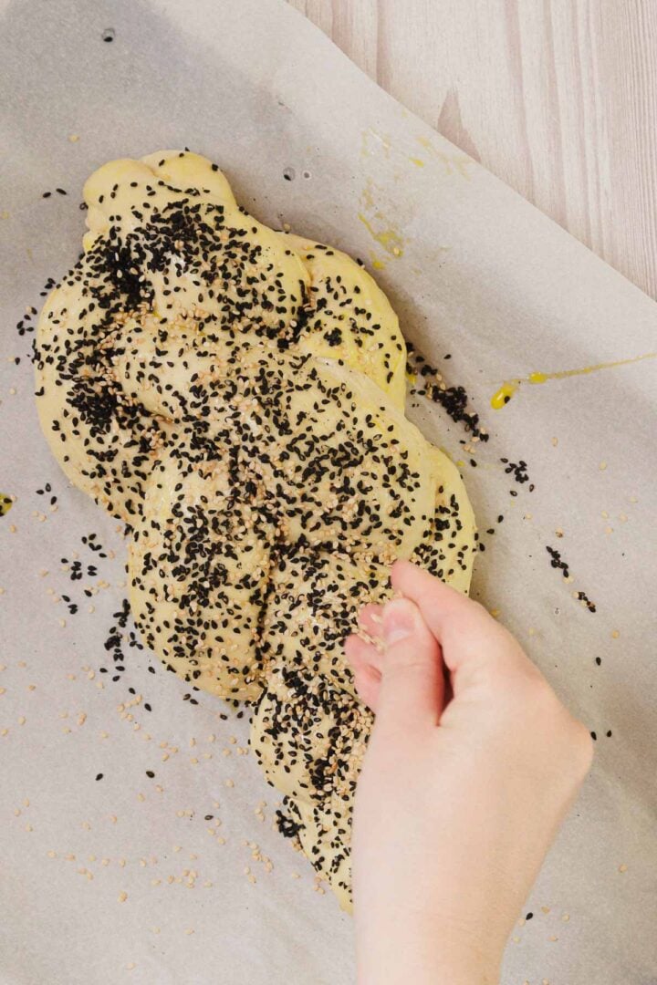 A hand sprinkles seeds onto Easy Challah braided bread dough on parchment paper before baking.