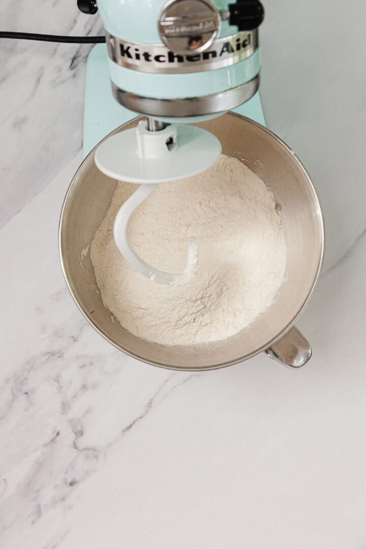 A stand mixer with a dough hook attachment and flour in the mixing bowl on a marble countertop, ready for Easy Challah.