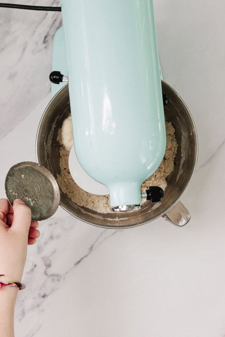 A hand adds an ingredient to a stand mixer with flour and dough inside, seen from above on a marble counter—capturing the simple steps of making Easy Challah.