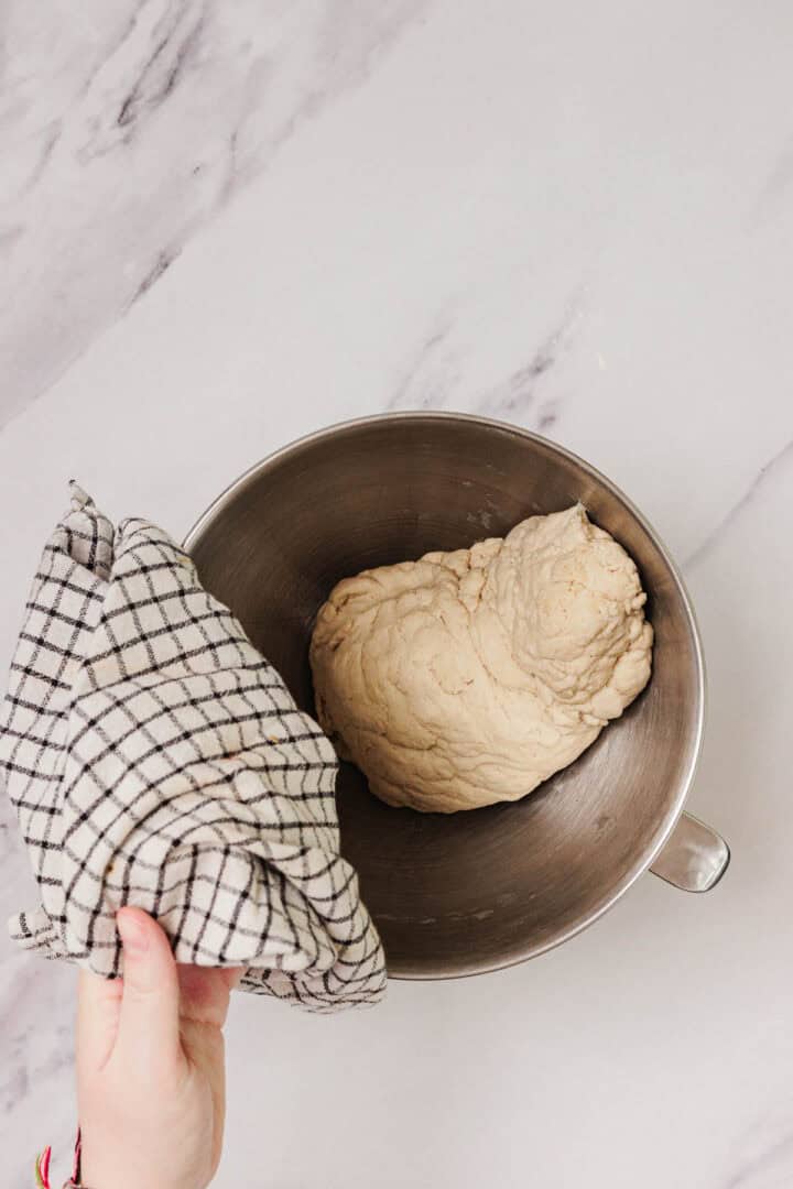 A hand holds a checkered towel over a metal bowl containing Easy Challah dough on a marble surface.