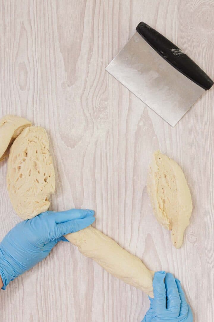 Gloved hands shaping Easy Challah dough on a light wooden surface, with a metal bench scraper nearby.