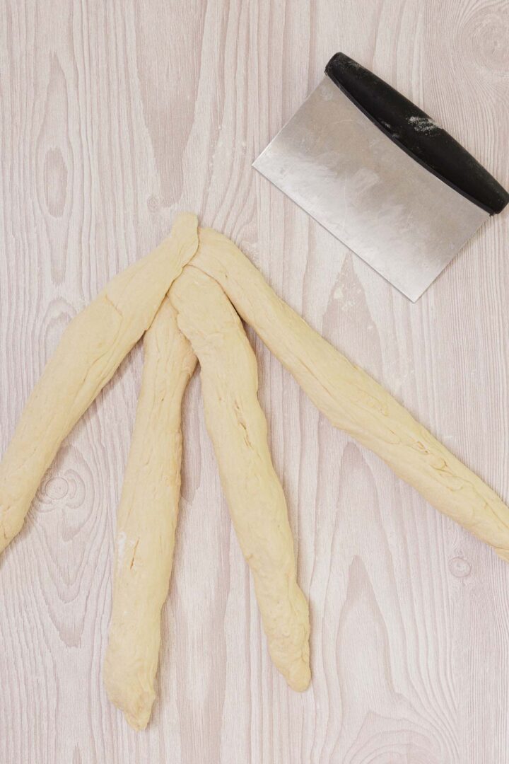 Three dough strands, ready for braiding Easy Challah, and a metal bench scraper on a light wooden surface.