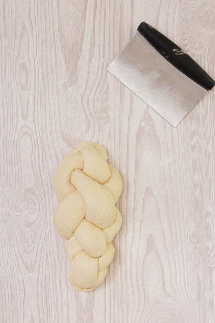 Easy Challah braided dough and a metal bench scraper rest on a light wood surface.