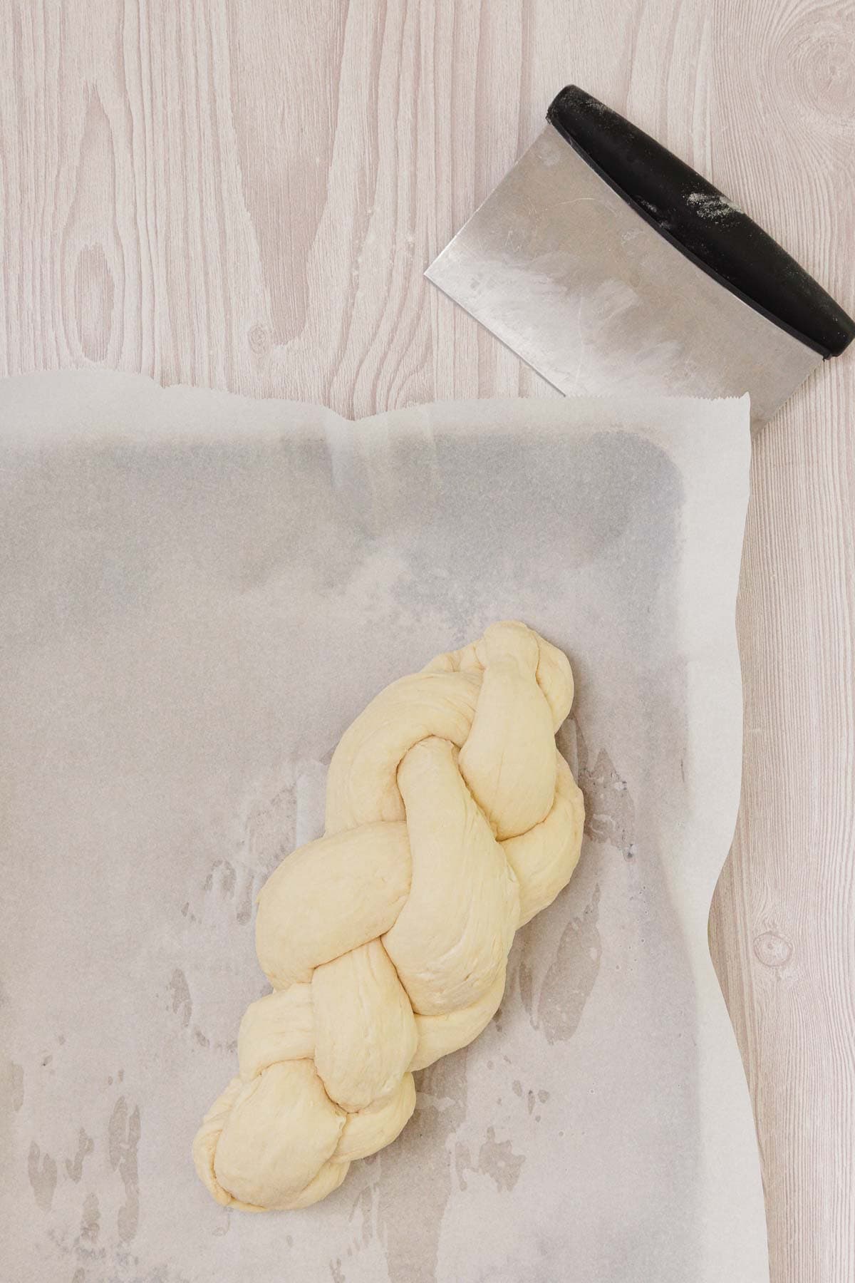 An easy Challah braided dough loaf rests on parchment paper with a dough scraper above it, set on a light wooden surface.