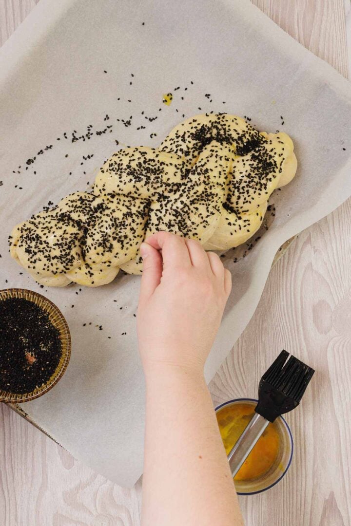 A hand sprinkles black seeds onto braided Easy Challah dough on parchment paper, next to a brush and egg wash.