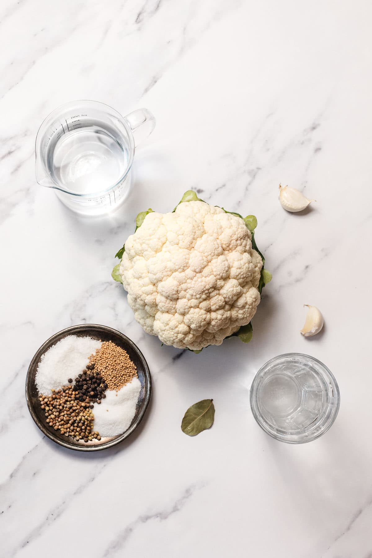 Overhead view of a cauliflower, spices, salt, garlic cloves, water in a jug, and a glass on a marble surface.