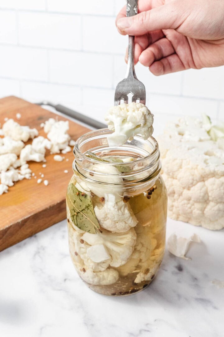 A hand lifts pickled cauliflower from a glass jar with a fork; chopped cauliflower on a cutting board nearby.