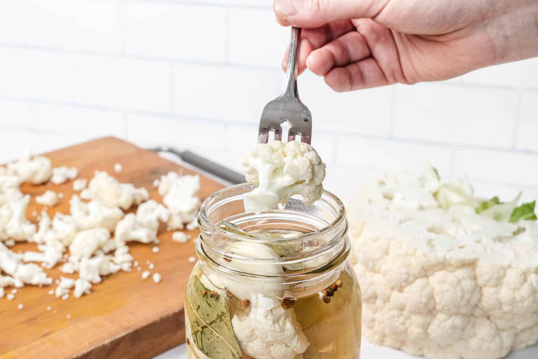 A hand uses a fork to lift pickled cauliflower from a glass jar; fresh cauliflower on a cutting board nearby.
