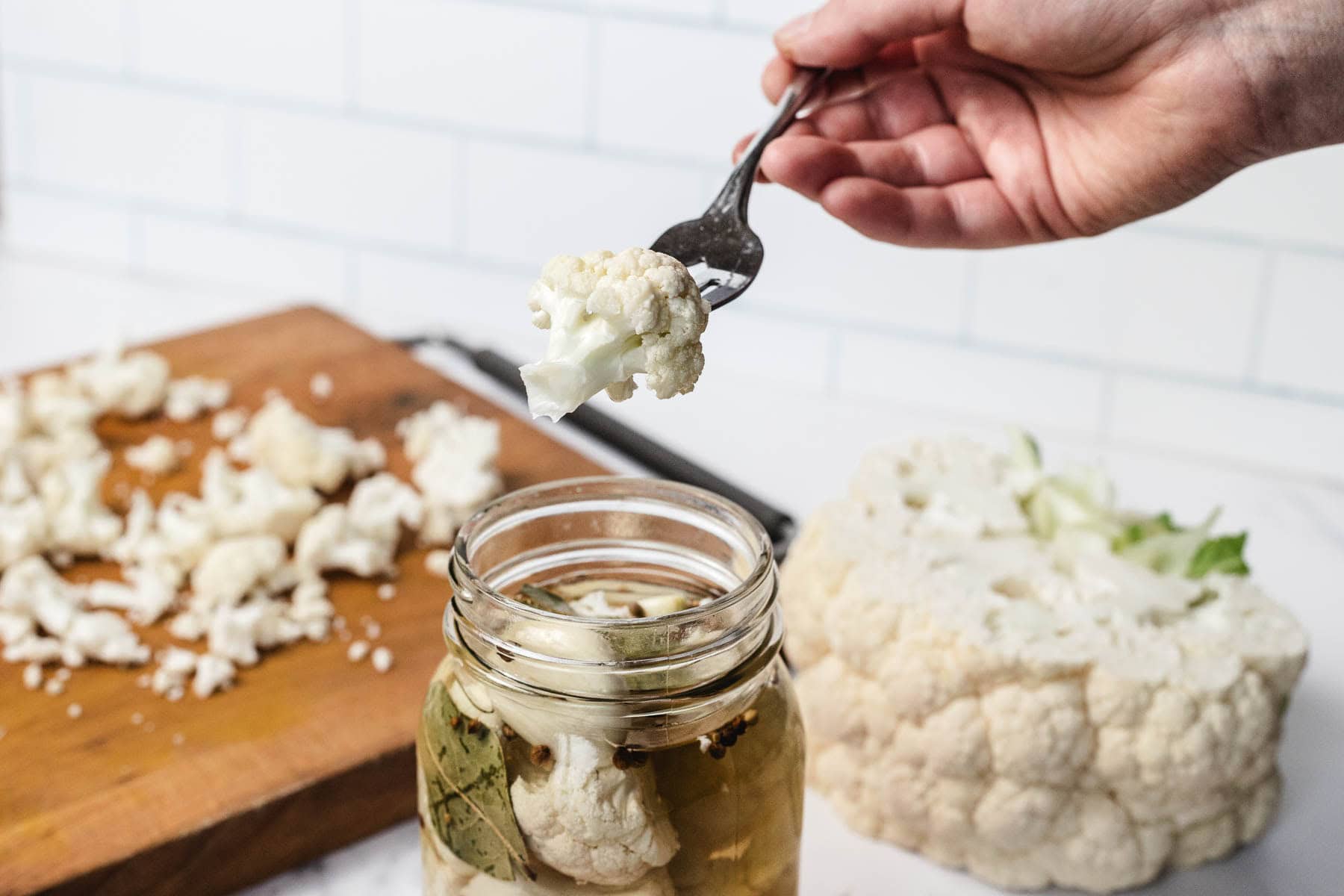 A hand holds a fork with pickled cauliflower over a jar, with a cutting board and fresh cauliflower nearby.