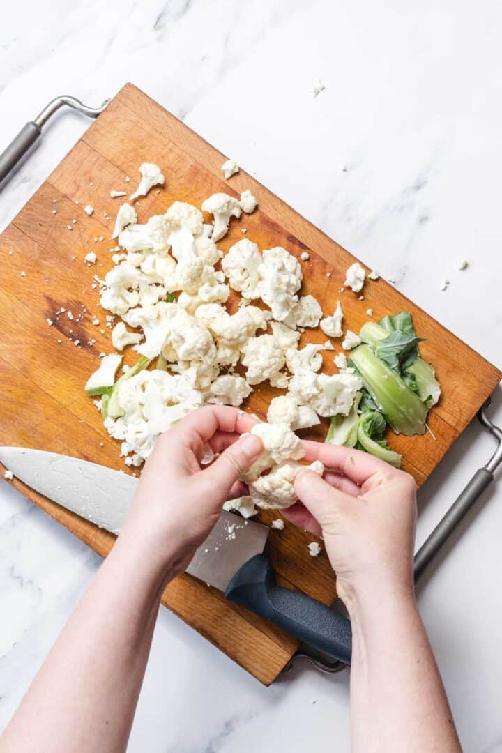 Hands breaking cauliflower florets on a wooden cutting board, with a knife and cauliflower stems nearby.