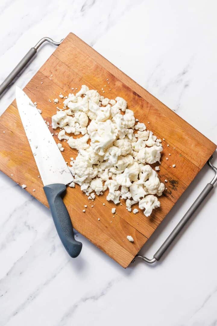 Chopped cauliflower and a kitchen knife on a wooden cutting board, placed on a marble counter.