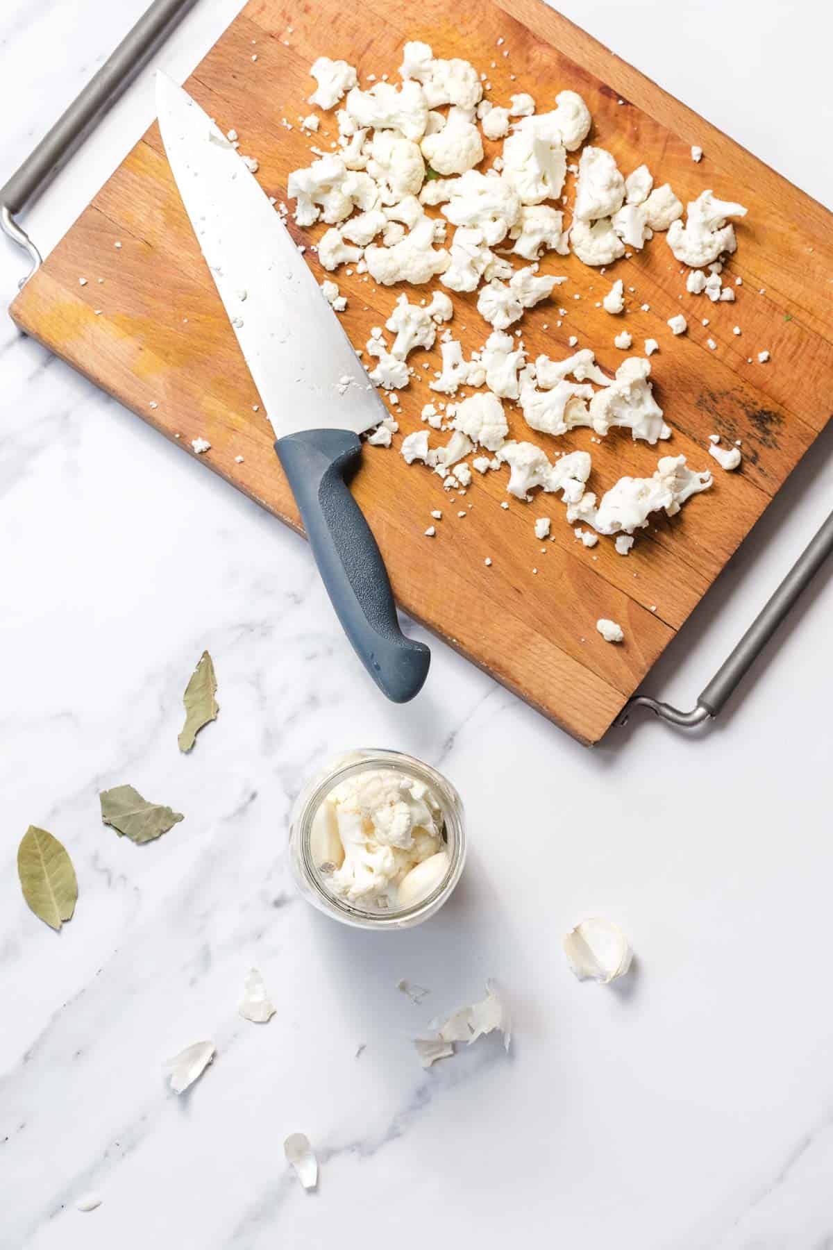 Chopped cauliflower and a knife on a cutting board, with bay leaves, garlic, and a jar of mayonnaise nearby.