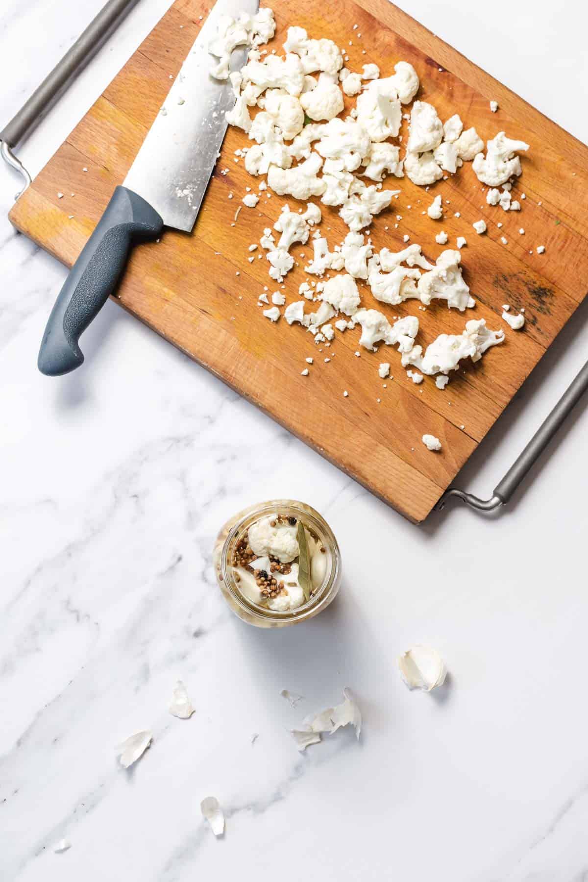 Chopped cauliflower on a wooden cutting board beside a knife and a jar with seasonings on a marble surface.
