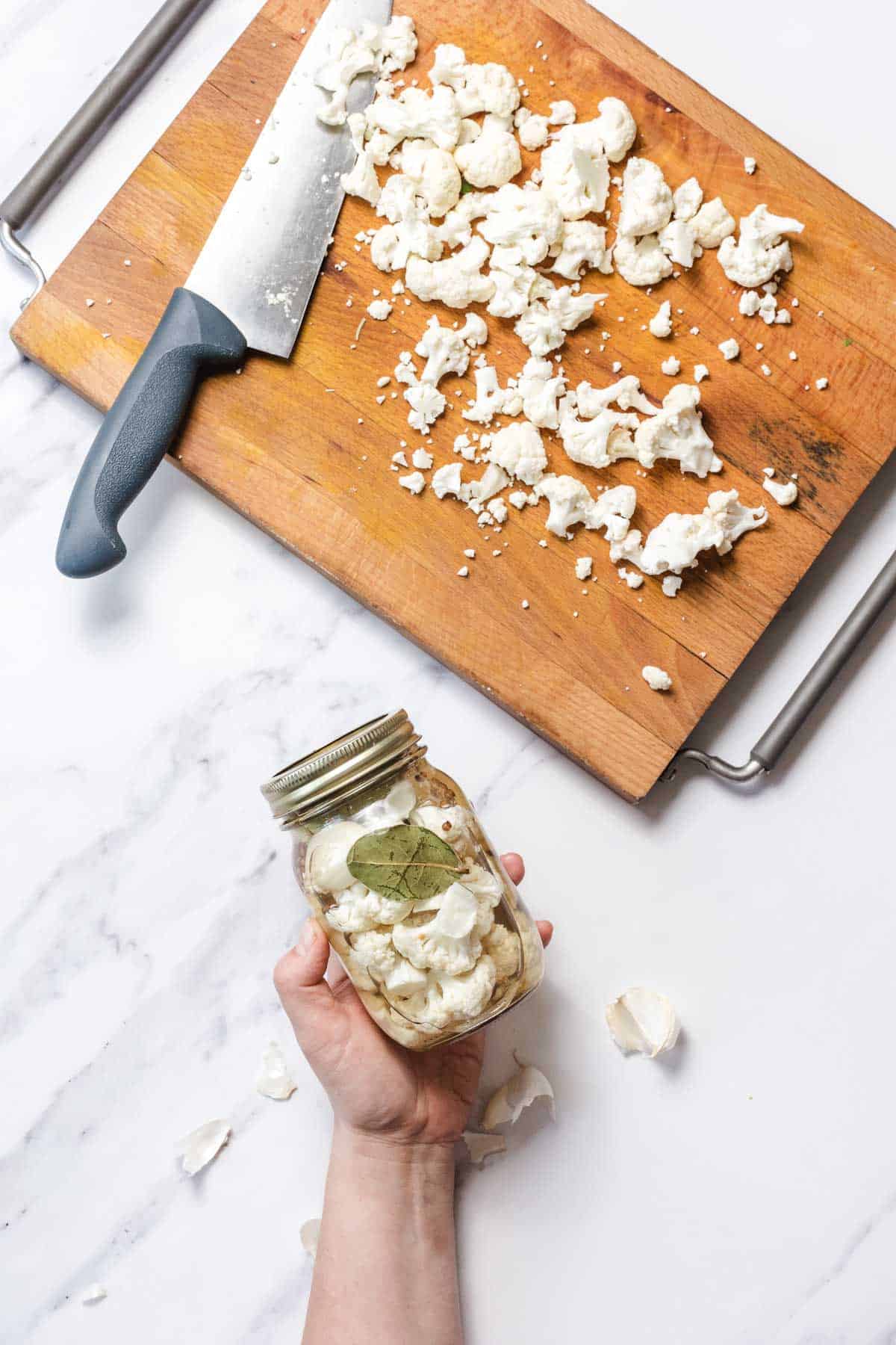 A hand holds a jar of pickled cauliflower next to a cutting board with chopped cauliflower and a knife.