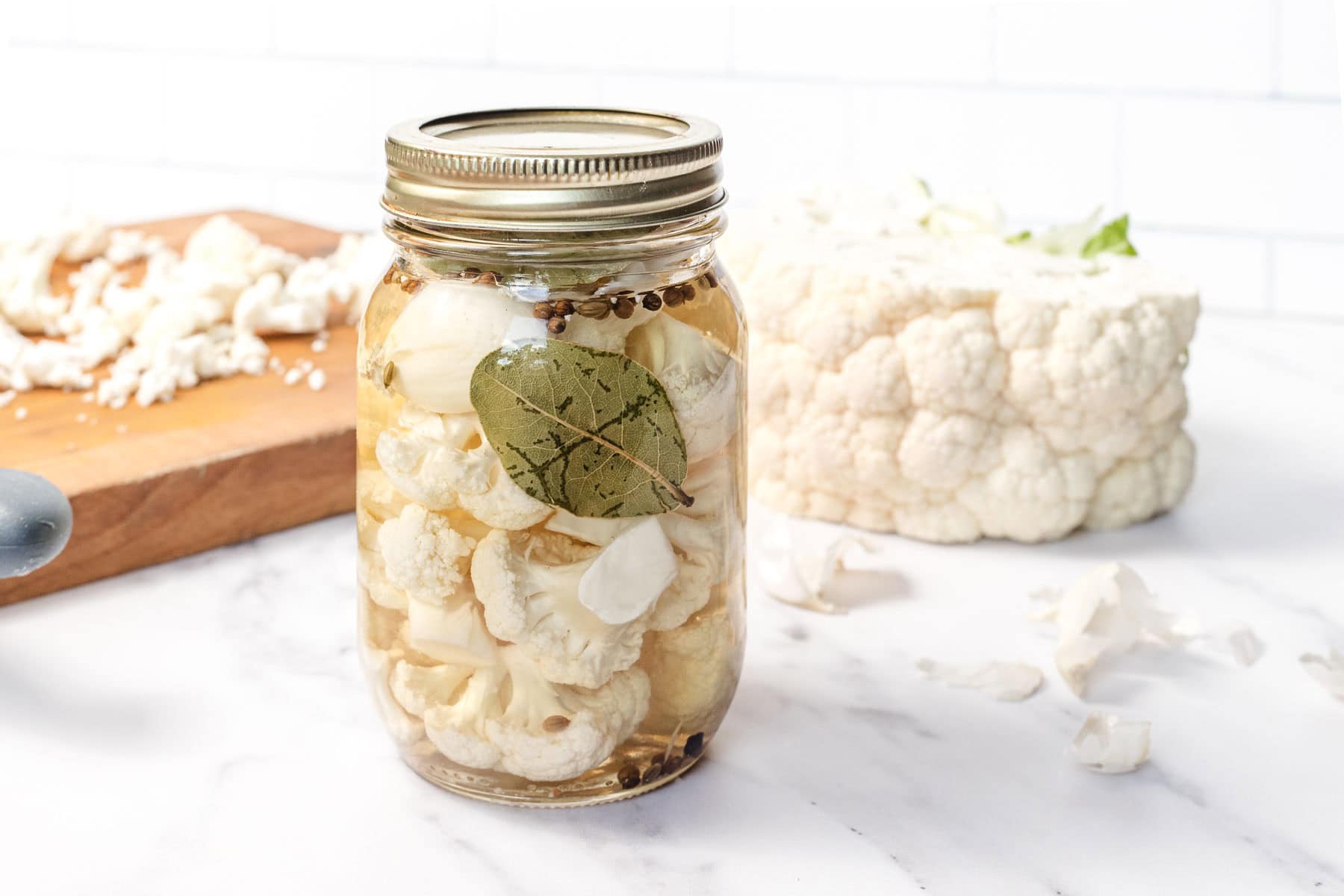 A jar of pickled cauliflower with garlic and bay leaf, next to fresh cauliflower on a white surface.