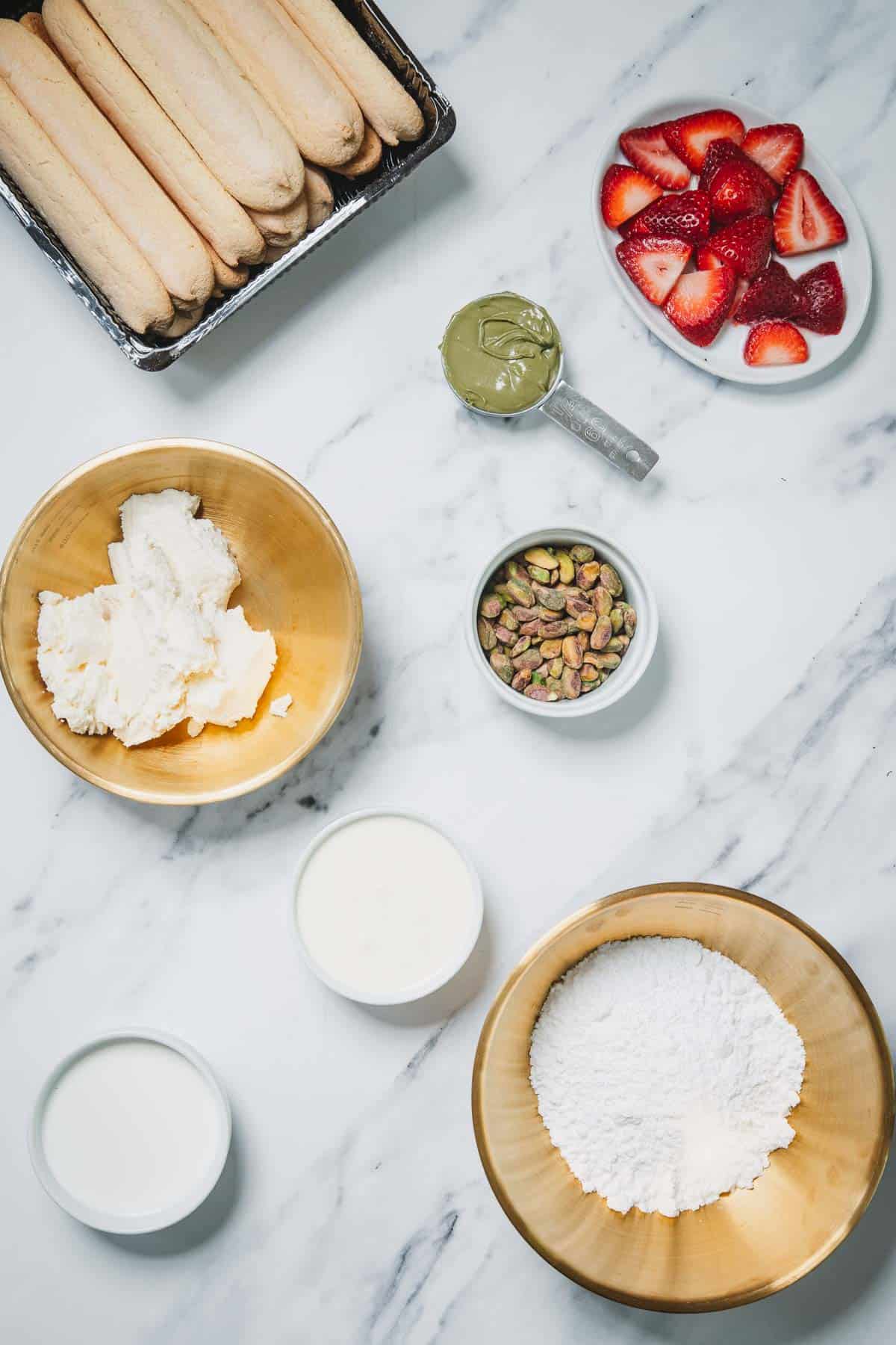 Ingredients on a marble counter for a No-Bake Strawberry Pistachio Ladyfingers Dessert: ladyfingers, strawberries, mascarpone, pistachios, matcha, cream, and sugar.