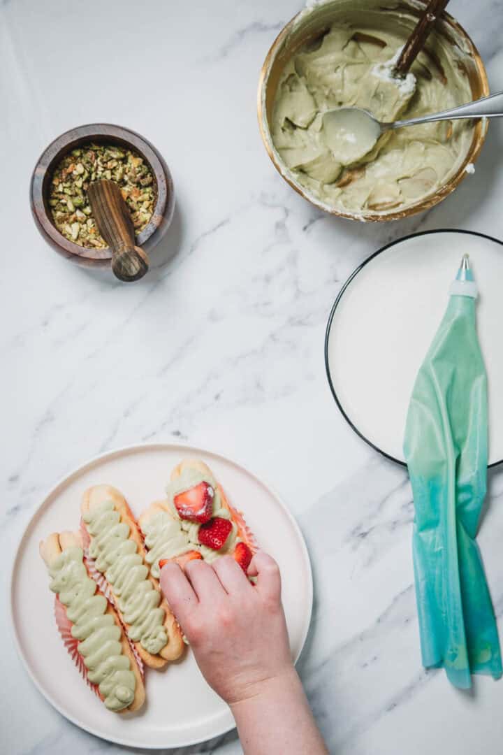 A hand places strawberries on pastries topped with green cream, creating a No-Bake Strawberry Pistachio Ladyfingers Dessert, with a piping bag and bowls nearby.