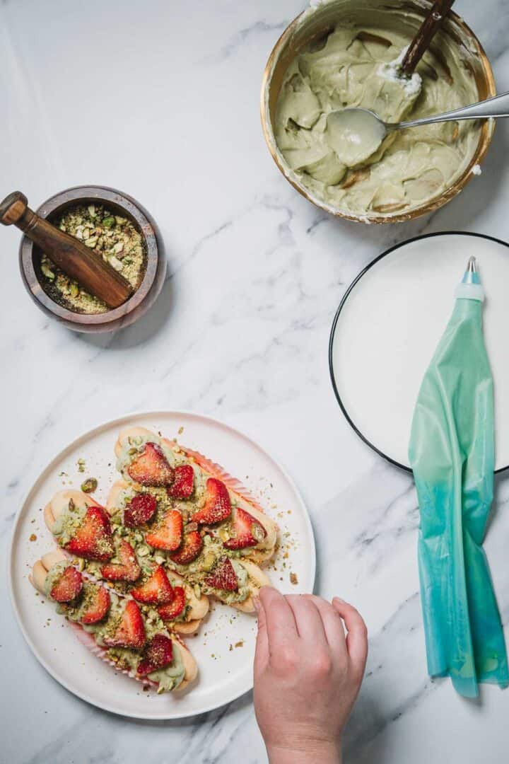 A hand decorates No-Bake Strawberry Pistachio Ladyfingers Dessert with crushed pistachios, beside bowls of cream and a piping bag.
